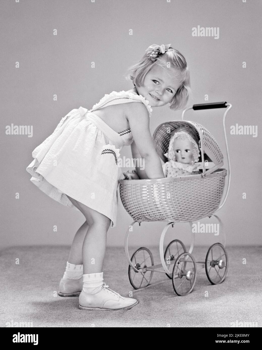 1940s SMILING BLONDE GIRL WEARING WHITE DRESS PICKING UP HER DOLL FROM A TOY STROLLER PRAM WHILE LOOKING OVER HER SHOULDER - j9292 HAR001 HARS B&W MATERNAL HAPPINESS CHEERFUL MODELS FEMININE SMILES CONCEPTUAL IMAGINATION JOYFUL ROLE-PLAYING STYLISH BABY BUGGY BEHAVIOR GENDER GROWTH JUVENILES STEREOTYPICAL BABY CARRIAGE BLACK AND WHITE CAUCASIAN ETHNICITY HAR001 OLD FASHIONED Stock Photo