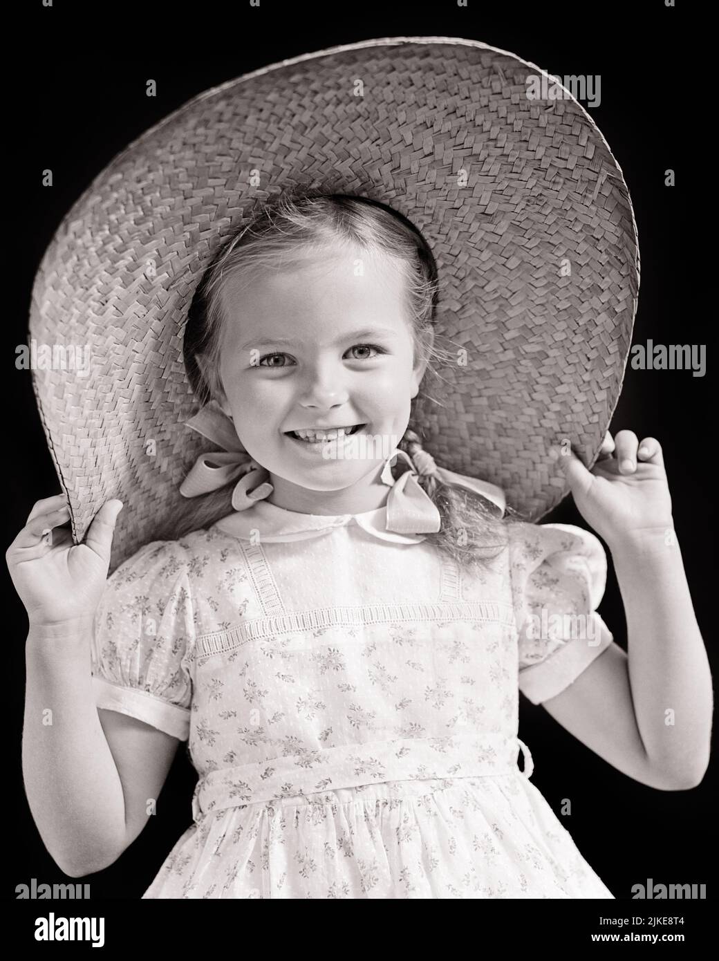 1940s SMILING PRETTY LITTLE GIRL WITH BRAIDS AND BOWS WEARING FLORAL PRINT DRESS HOLDING ON BIG STRAW HAT AND LOOKING AT CAMERA - j9996 HAR001 HARS COMMUNICATION PLEASED JOY LIFESTYLE FEMALES FLORAL STUDIO SHOT HEALTHINESS HOME LIFE COPY SPACE HALF-LENGTH CONFIDENCE EXPRESSIONS B&W SUMMERTIME PRETTY SUNSHINE HAPPINESS CHEERFUL AND EXCITEMENT PRIDE SMILES BOWS JOYFUL STYLISH BRAIDS CHARMING JUVENILES BLACK AND WHITE CAUCASIAN ETHNICITY HAR001 OLD FASHIONED Stock Photo