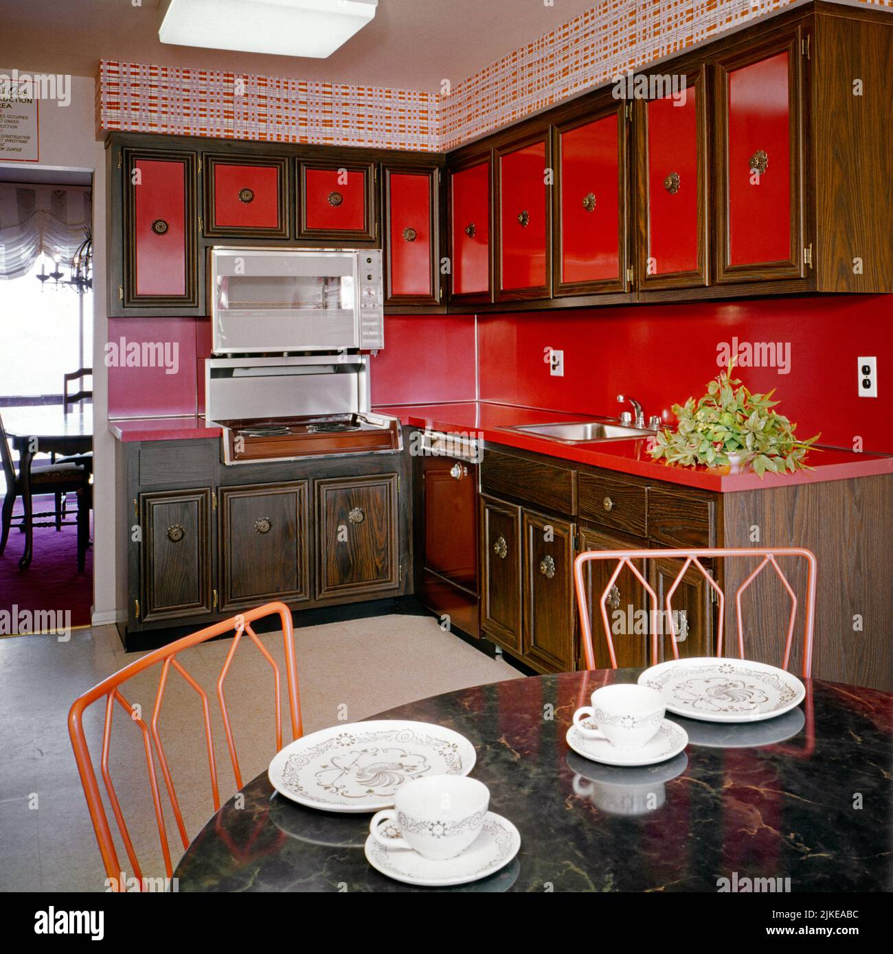 1970s EAT-IN KITCHEN WITH BRIGHT RED COUNTER TOPS CABINET FRONTS AND WALL PAPER DARK WOOD FRAMES AND FLORAL MURAL BY TABLE - ki2160 HAR001 HARS MURAL OLD FASHIONED Stock Photo