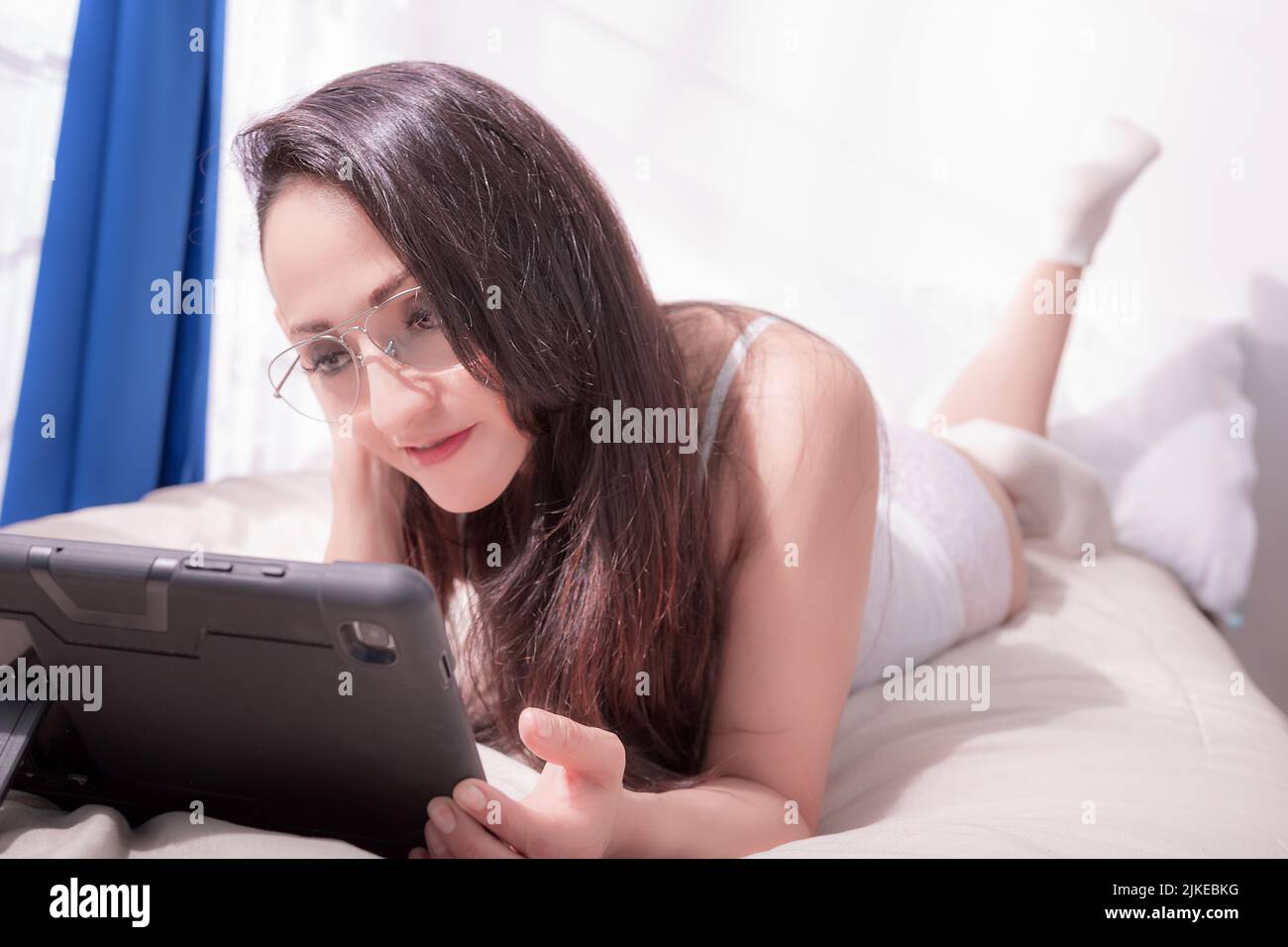 Beautiful woman in white underwear is very comfortable on the bed of her room where a lot of sunlight enters, looking at her electronic tablet in a ve Stock Photo