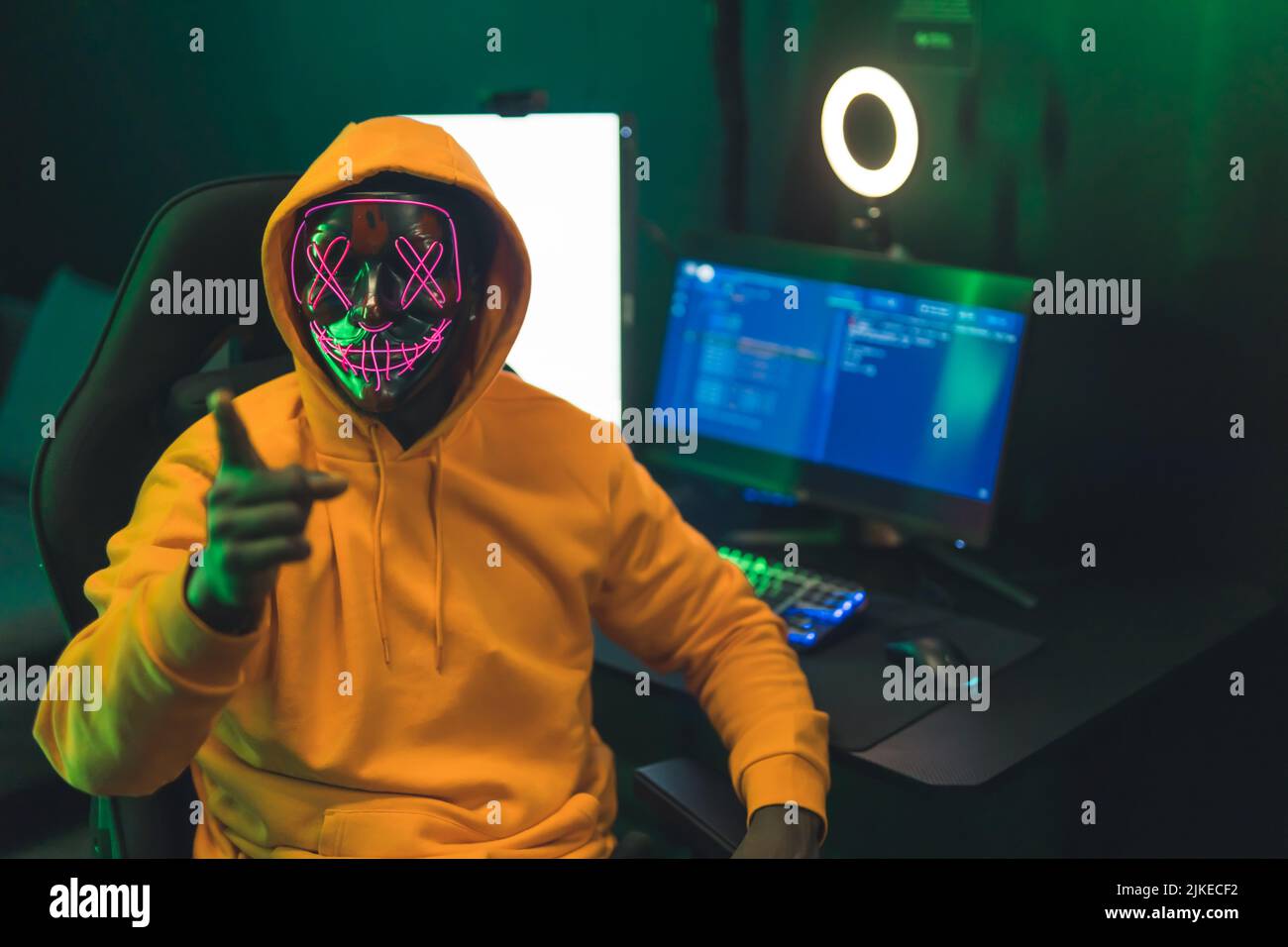 Young black man in orange hoodie and strange mask pointing at camera sitting by desk with gaming setup and ring light. Dark green lighting. High quality photo Stock Photo