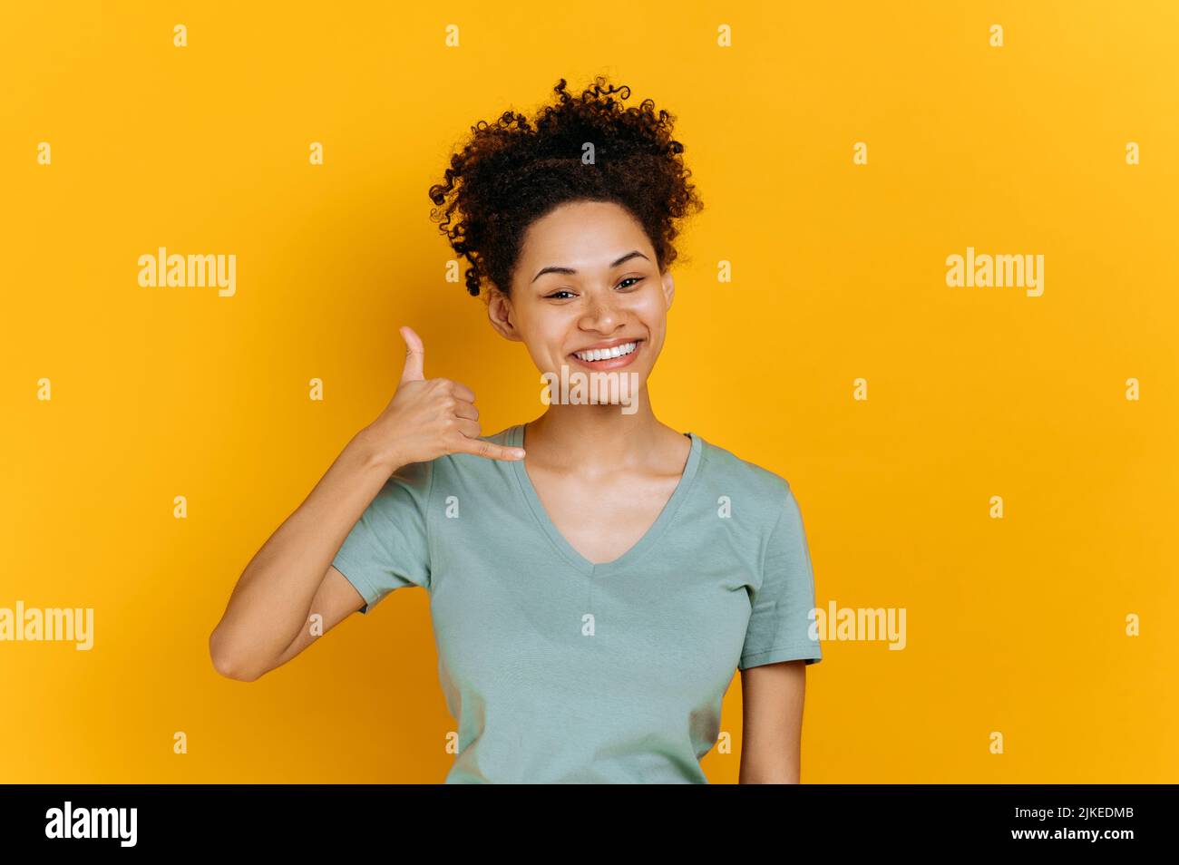 Call me. Positive african american millennial girl, holding hand near ear, showing telephone sign with fingers, asking for number, standing on isolated orange background, looks at camera, smiling Stock Photo