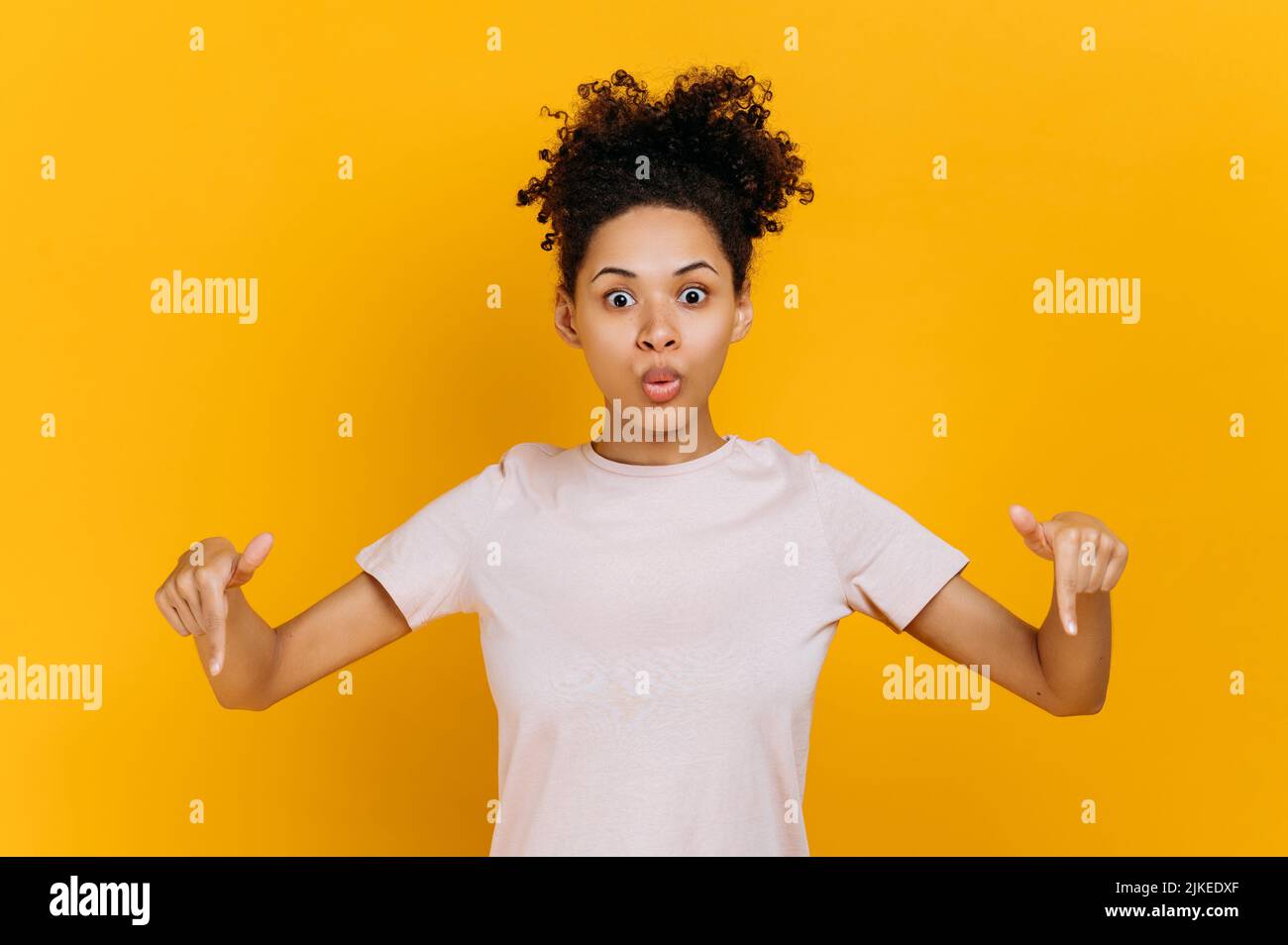 Shocked confused african american curly haired young woman, surprised looks at the camera and points fingers down, at empty copy space, stands on isolated orange color background Stock Photo