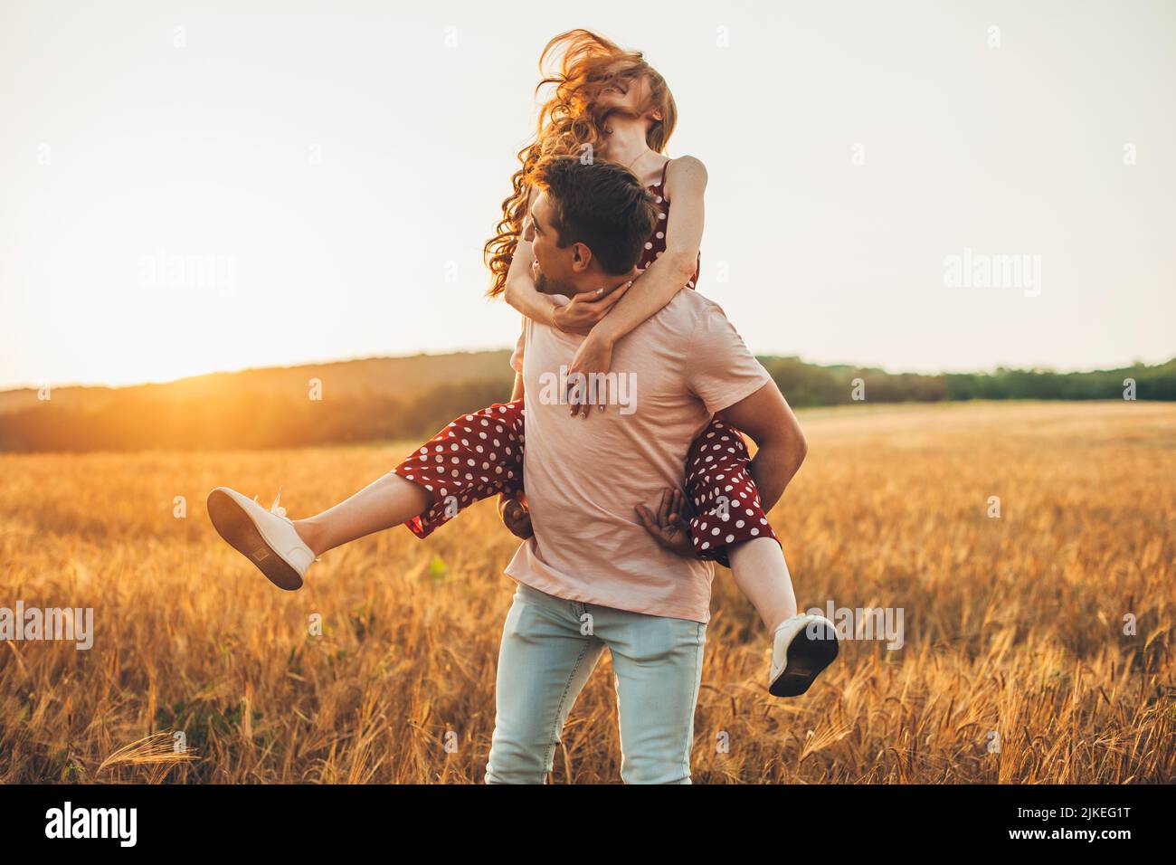 Attractive caucasian couple enjoying a day in a colorful yellow wheat field, man giving the girl a piggy back ride as they smile. Good day, happiness Stock Photo