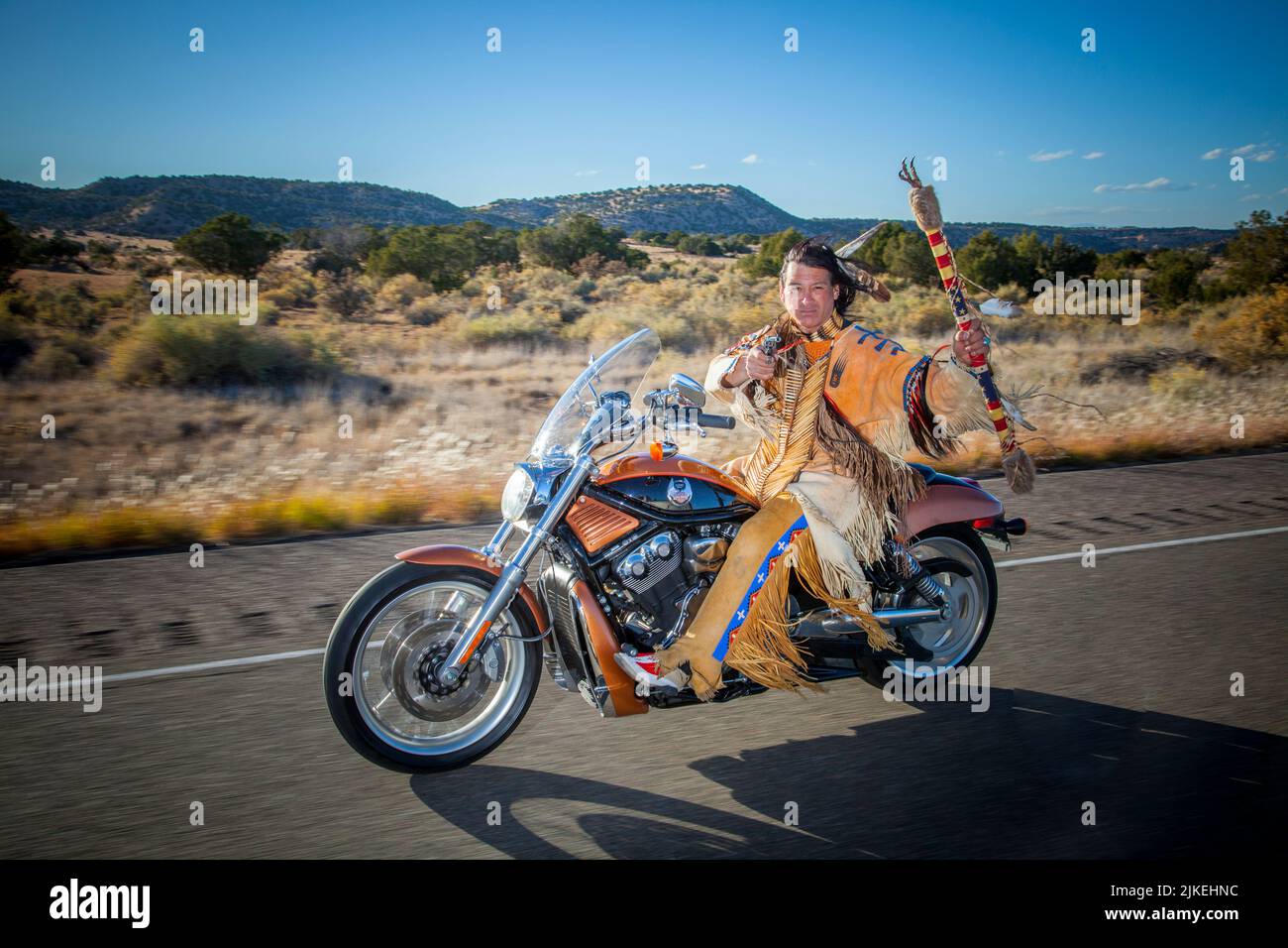 Traditionally dressed Arapahoe man holds up coup stick and gun while riding a Harley Davidson motorcycle down country highway Stock Photo