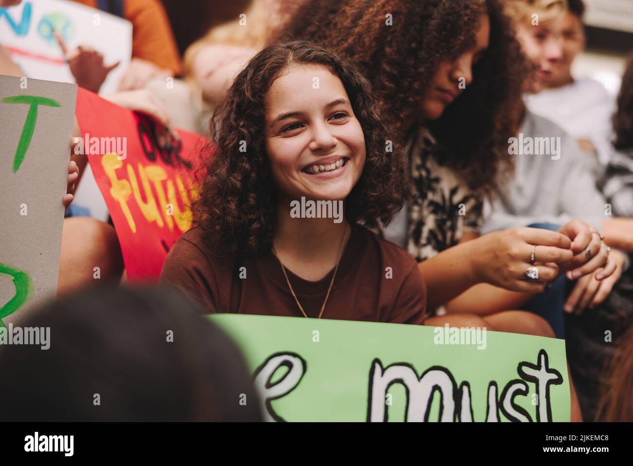 Happy teenage girl smiling cheerfully while sitting with a group of climate activists. Multicultural protestors striking for climate justice and envir Stock Photo