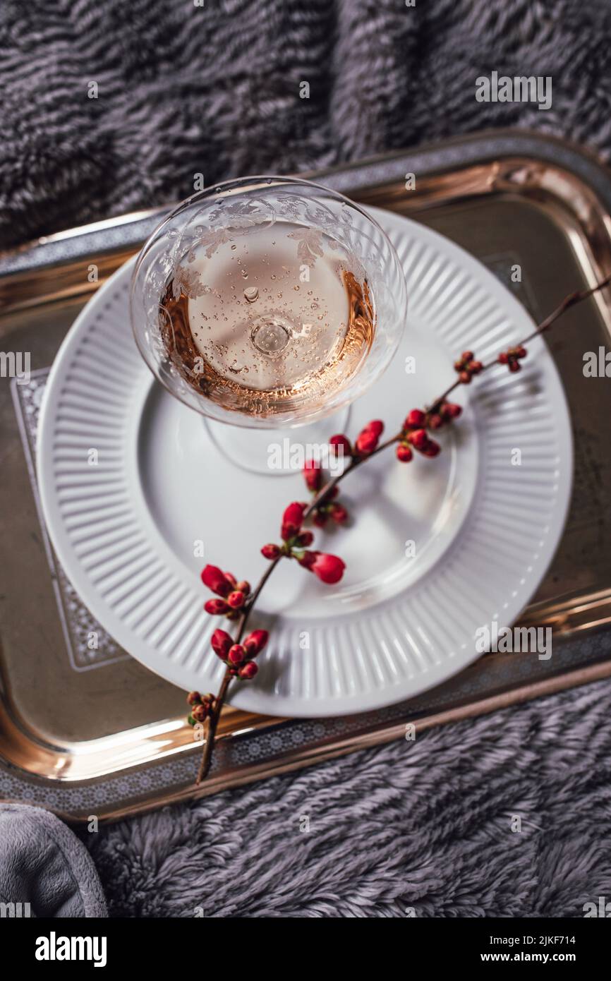 close up of not yet bloomed pink japanese flowering quince branch on white plate with rose wine, silver tray, gray faux fur blanket Stock Photo