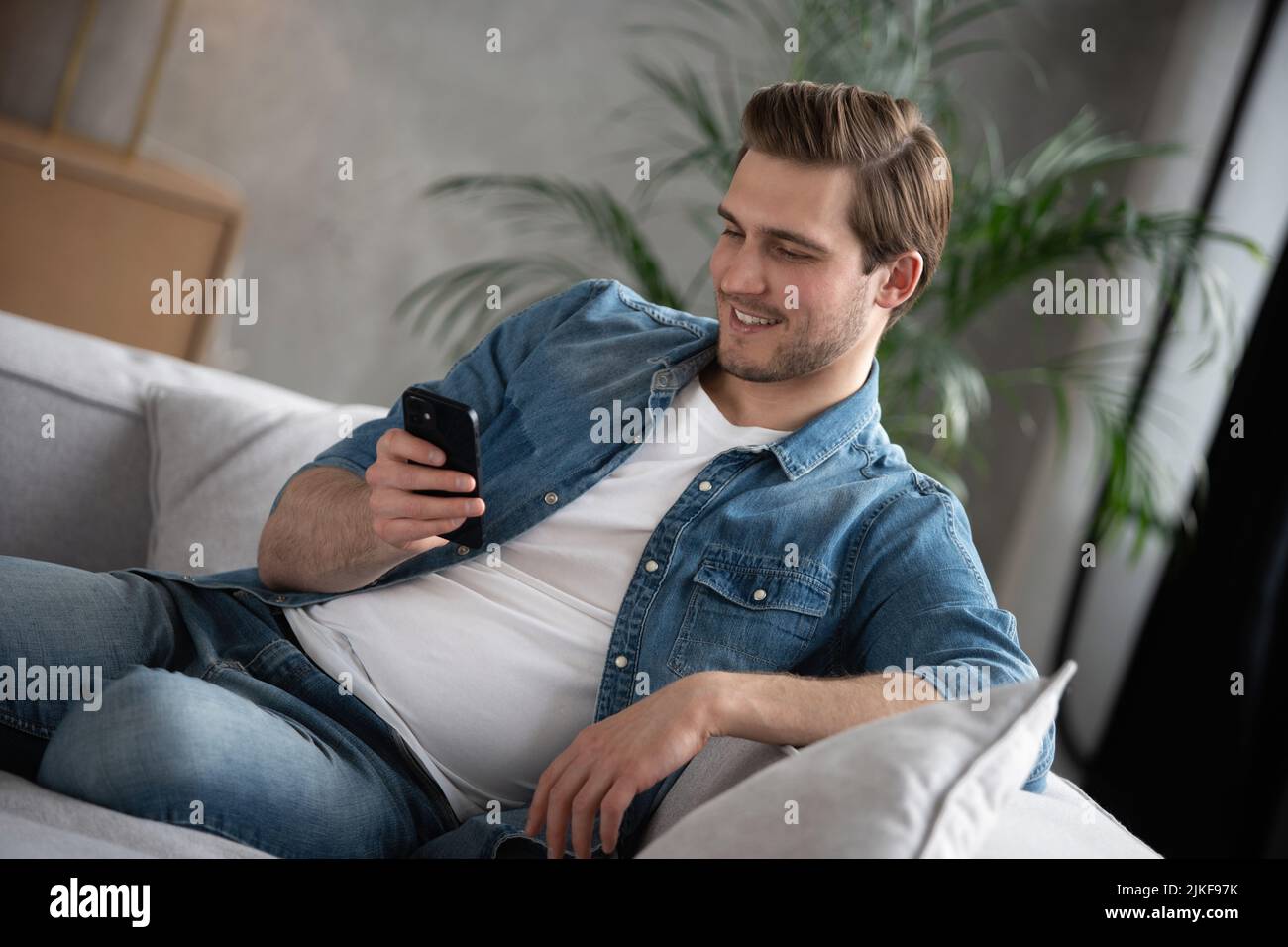 Attractive smiling young man wearing casual clothes sitting on a couch at the living room, using mobile phone Stock Photo