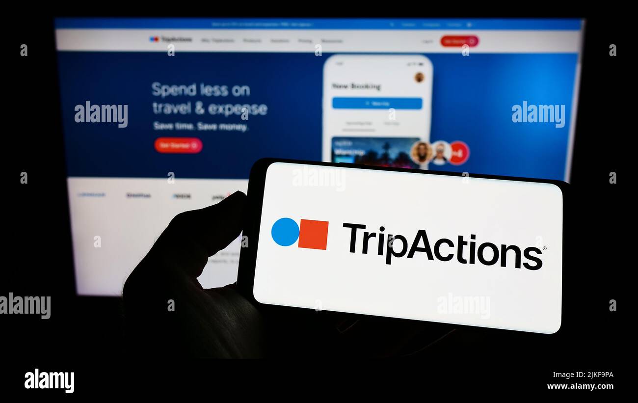 Person holding cellphone with logo of US travel management company TripActions Inc. on screen in front of web page. Focus on phone display. Stock Photo
