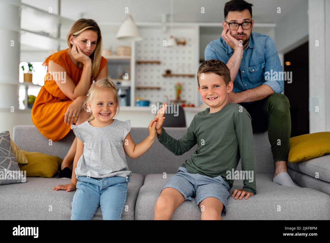 Tired parents sitting on couch feels annoyed exhausted while happy children playing together. Stock Photo