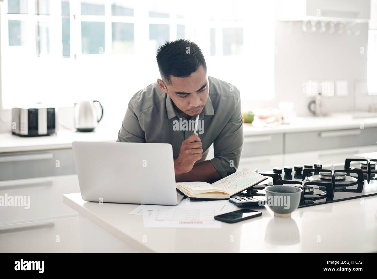 Going through his notes very carefully. a young man using a laptop while going through notes in the kitchen at home. Stock Photo