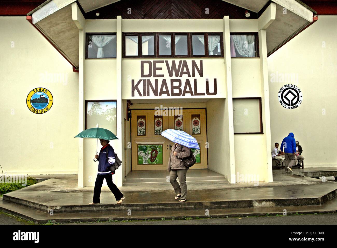 People carrying umbrellas as they are walking in front of the building of Dewan Kinabalu (Kinabalu Hall) in Kinabalu Park, Ranau, Sabah, Malaysia. Stock Photo