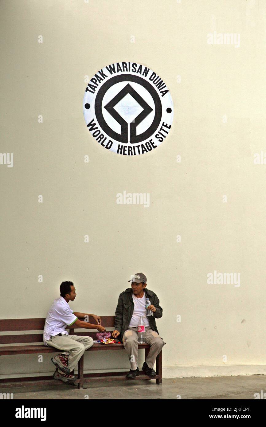 Men having a conversation as they are sitting on a bench in front of a wall decorated with 'World Heritage' sign at the building of Dewan Kinabalu (Kinabalu Hall) in Kinabalu Park, Ranau, Sabah, Malaysia. Stock Photo