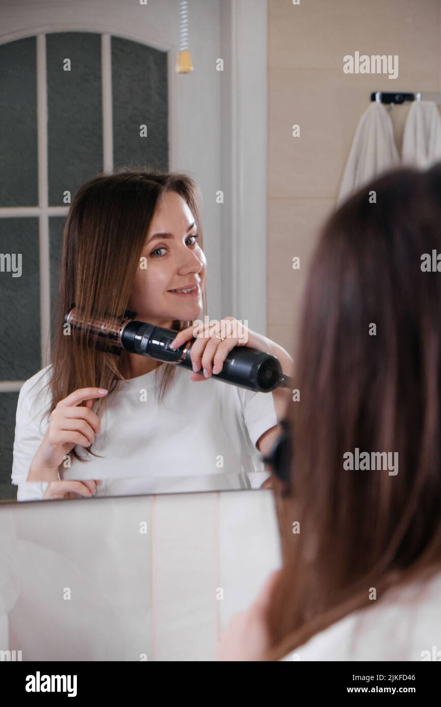 Woman drying her hair, styling in the bathroom, looking at the mirror, combing hair at home Stock Photo