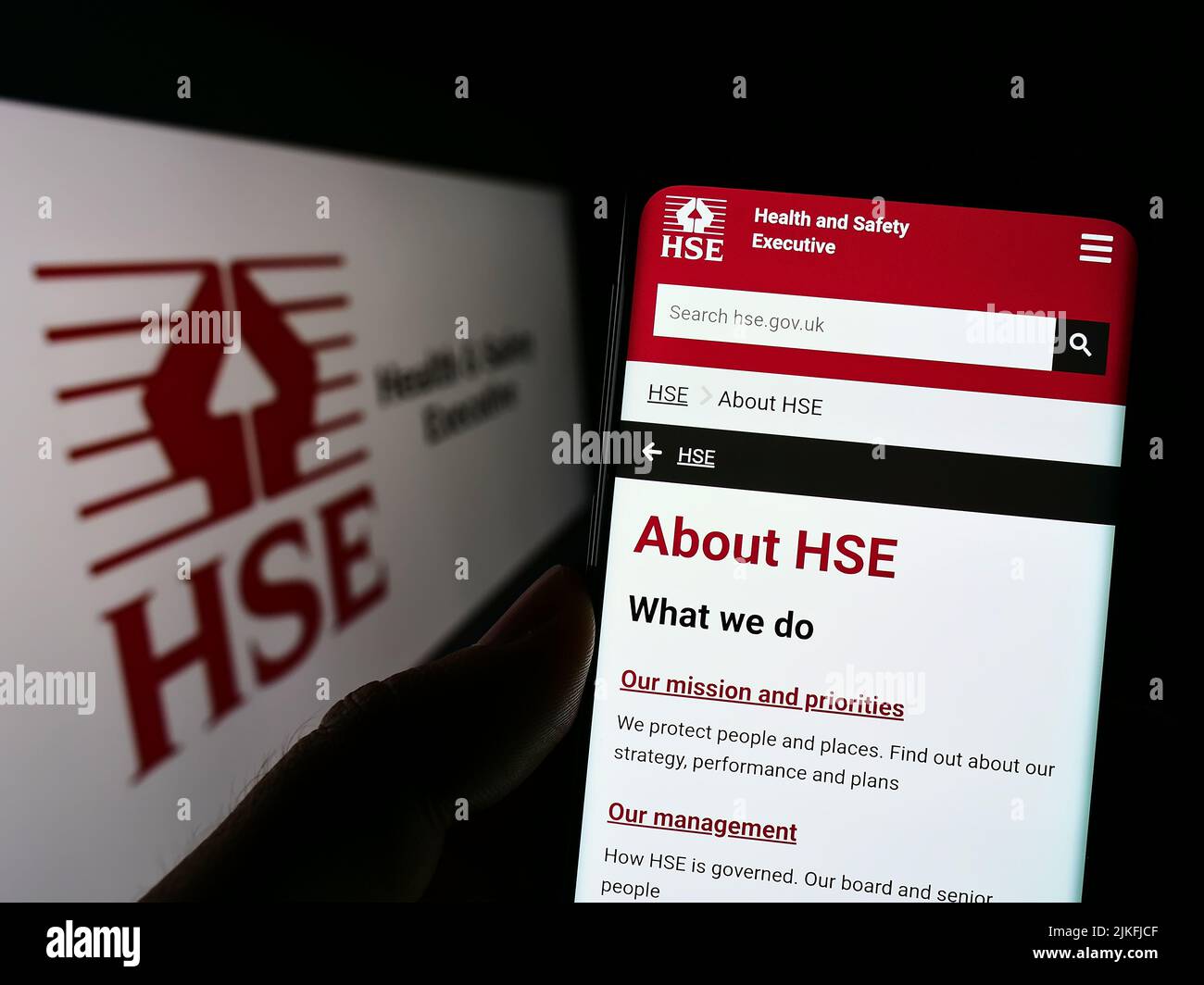 Person holding cellphone with website of UK agency Health and Safety Executive (HSE) on screen in front of logo. Focus on center of phone display. Stock Photo