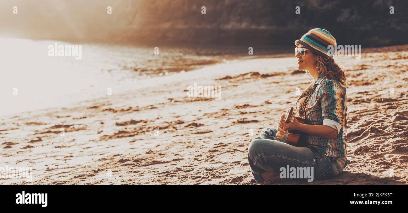 Female tourist enjoy summer holiday vacation sitting at the beach during sunset playing a guitar alone. Concept of happy life and travel destination. Stock Photo