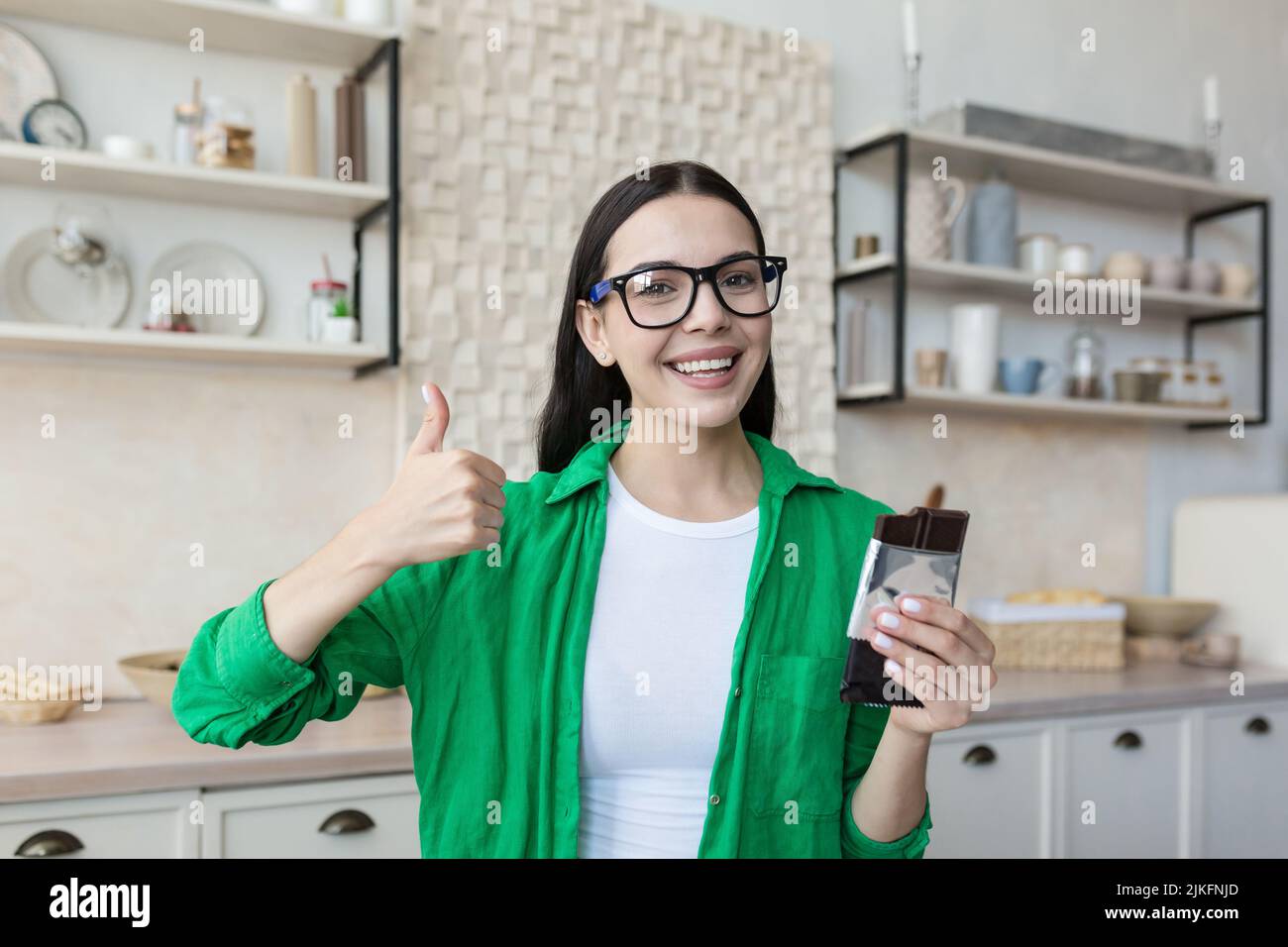 Young beautiful woman teenager holding chocolate bar in hands in glasses and green shirt, looking at camera smiling, recommending holding thumb up Stock Photo
