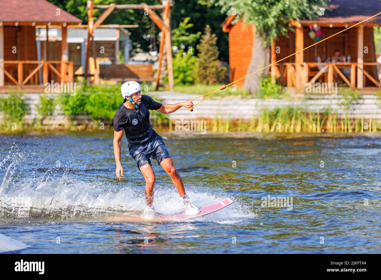 A wakeboarder in a white safety helmet rides a water board in a river bay on a summer day. 06.19.1922. Kyiv. Ukraine. Stock Photo