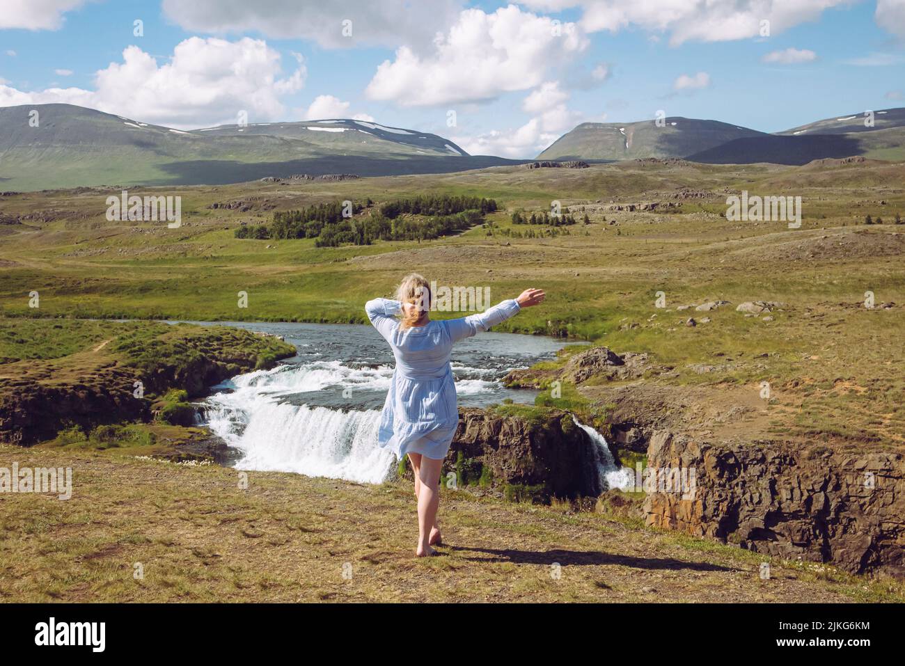 Woman wearing blue summer dress in wild nature in Iceland, Europe. Green mountains and waterfall on background, blue sky, sunny day. Stock Photo