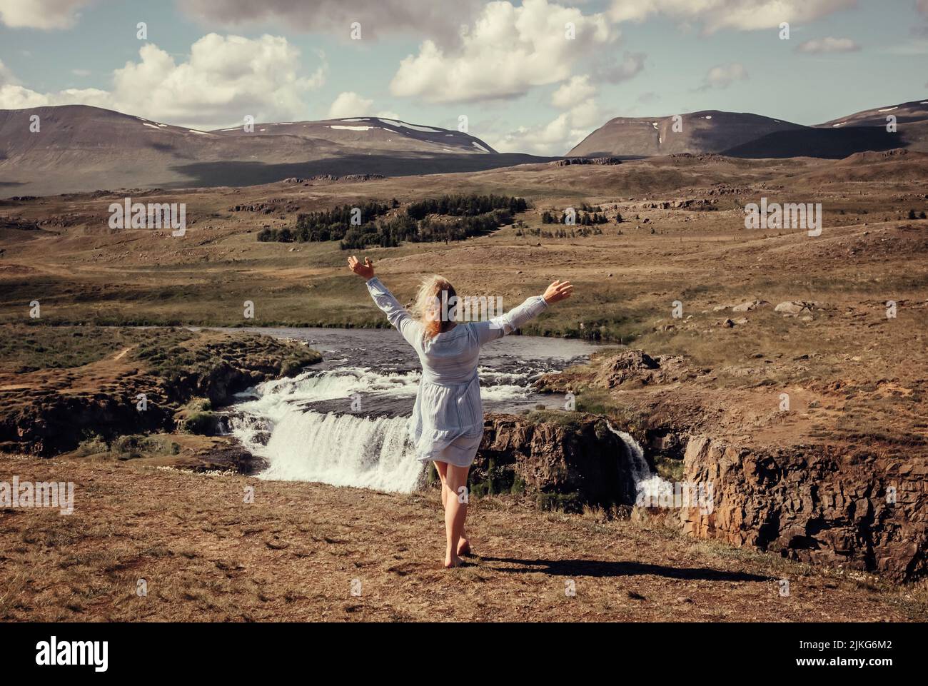 Woman wearing blue summer dress in wild nature in Iceland, Europe. Green mountains and waterfall on background, blue sky, sunny day. Stock Photo