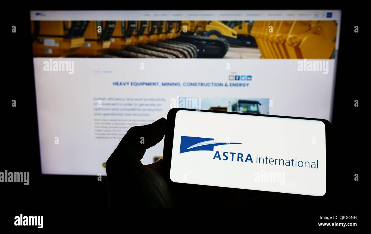 Person holding cellphone with logo of Indonesian company PT Astra International Tbk on screen in front of webpage. Focus on phone display. Stock Photo