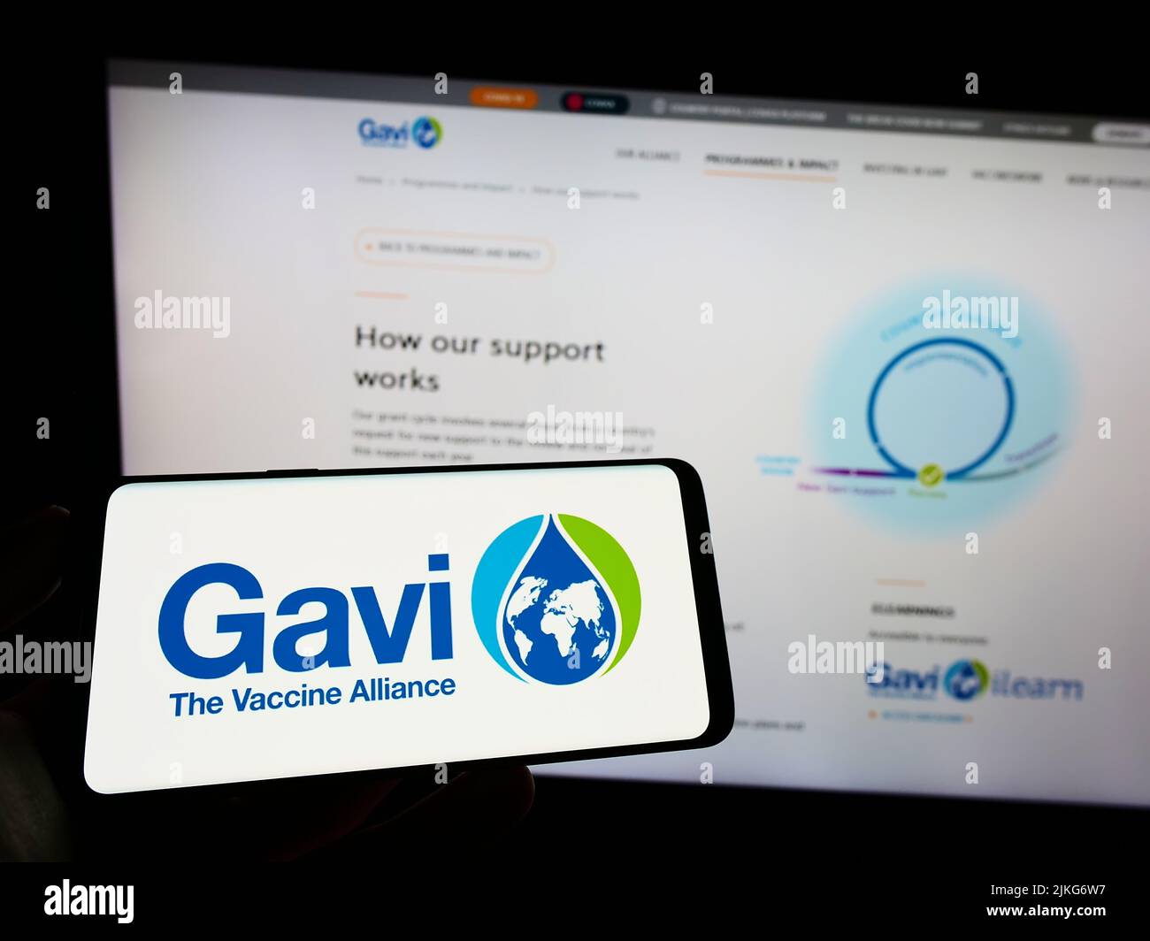 Person holding cellphone with logo of health partnership Gavi, the Vaccine Alliance on screen in front of webpage. Focus on phone display. Stock Photo