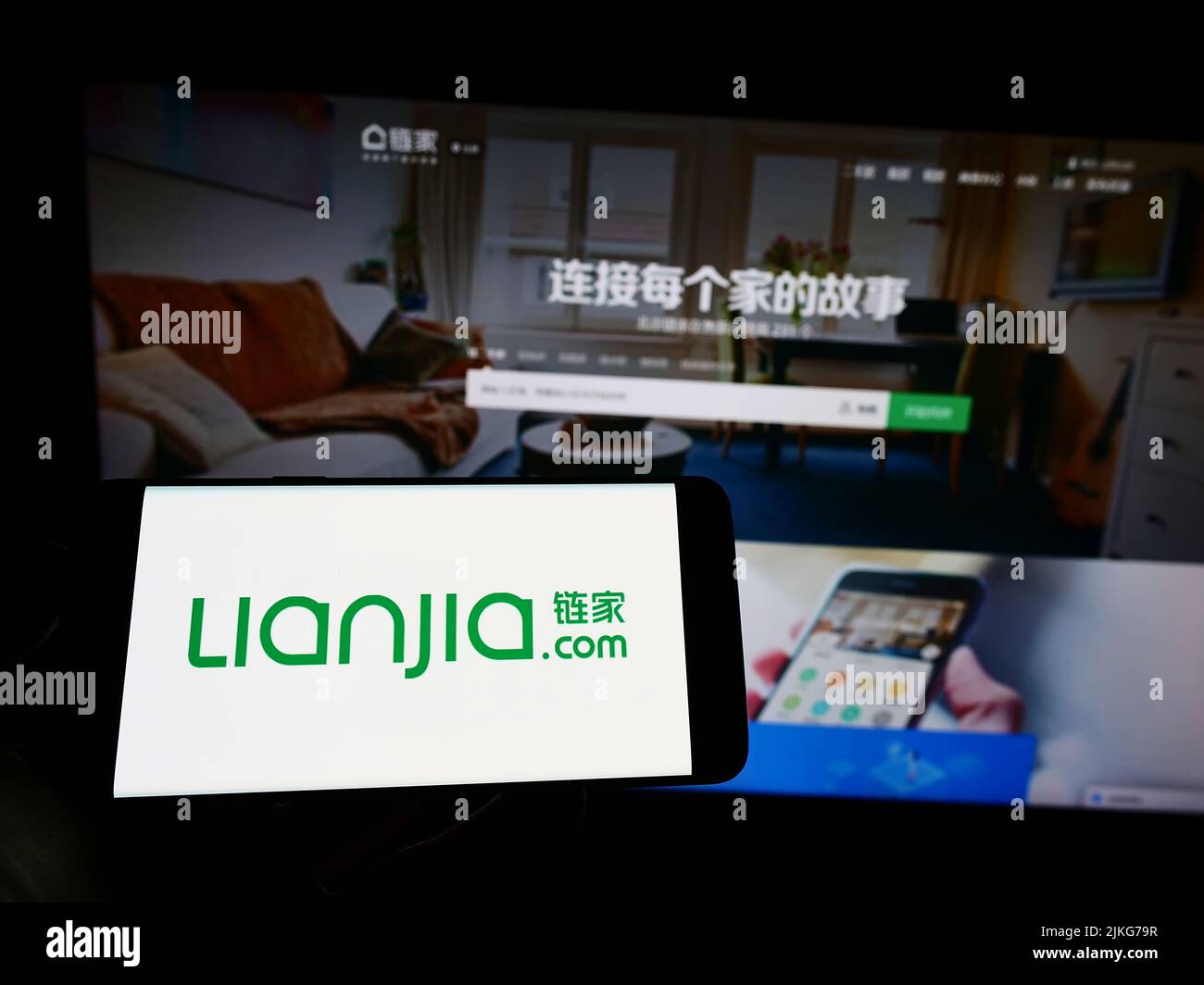 Person holding mobile phone with logo of Chinese real estate company Lianjia on screen in front of business web page. Focus on phone display. Stock Photo