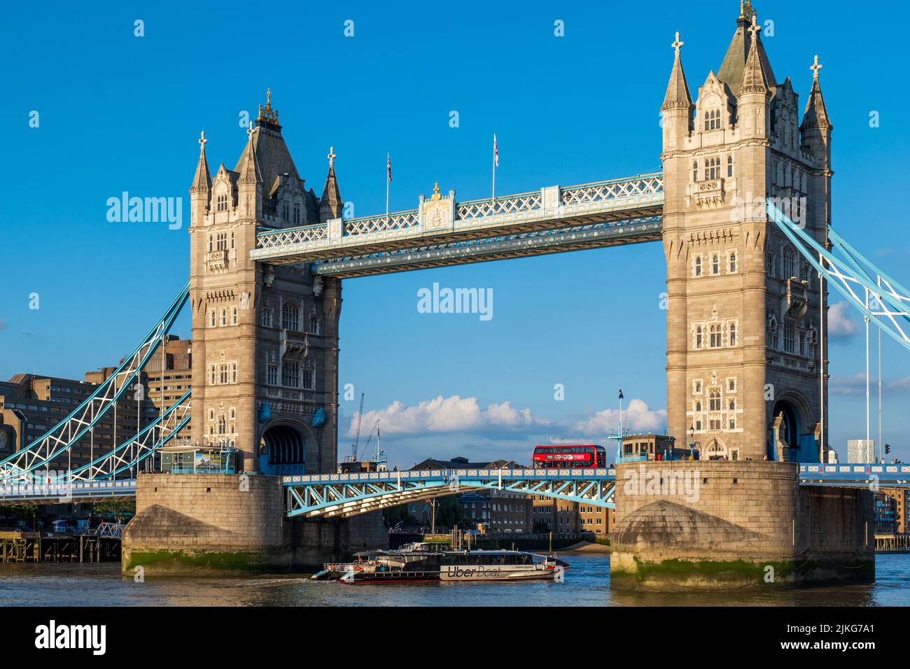 An Uber Boat by Thames Clippers on the River Thames passes beneath Tower Bridge whilst an iconic red London bus crosses the bridge above. Stock Photo