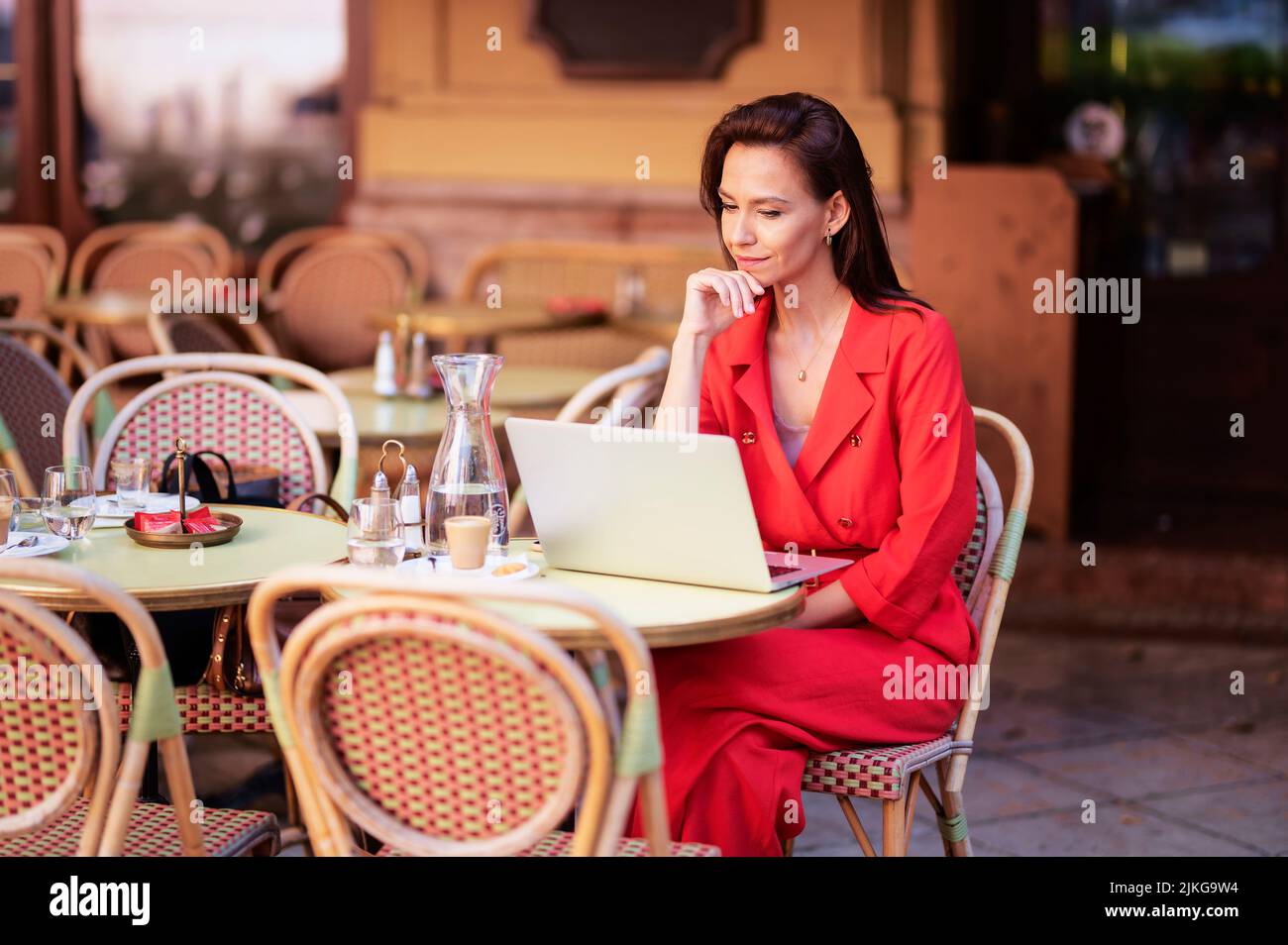Attractive woman looking thoughtfully and using a laptop while sitting at outdoor cafe terrace. Stock Photo