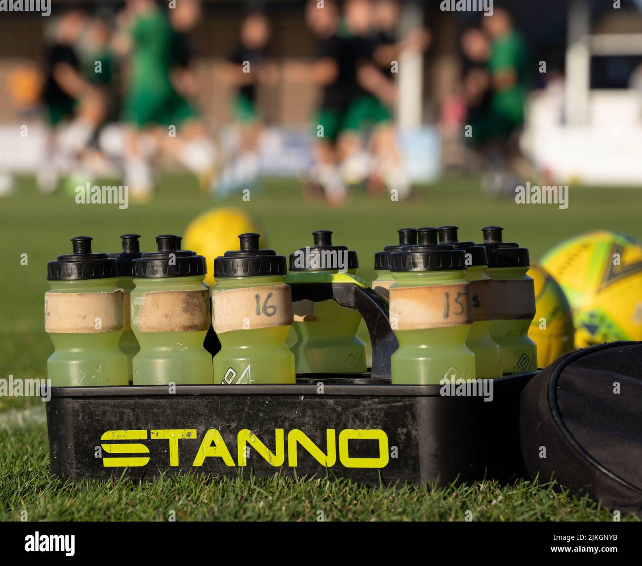 Yellow plastic refillable water bottles with black tops sit in a STANNO carrier on a grass football pitch (soccer). Players warm up in the background. Stock Photo