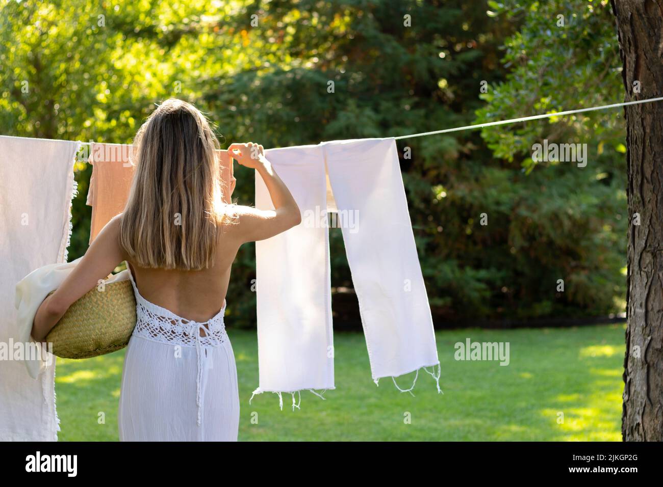 Back view of unrecognizable young blonde woman on boho outfit hanging up the washing in the garden in summer time with copy space Stock Photo