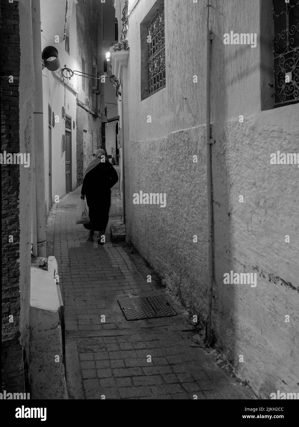 A vertical grayscale shot of a female walking through an alley in Tangier, Morocco Stock Photo