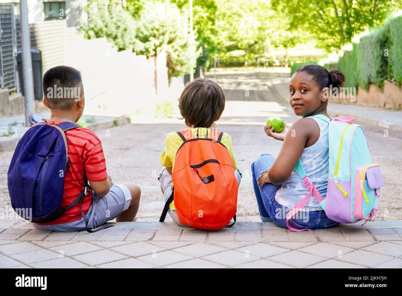 multiethnic kids with backpacks sitting on the street at school entrance eating apples. Back to school concept. Multiethnic children group Stock Photo
