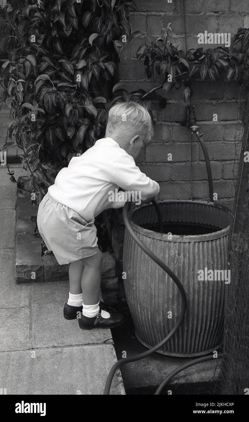 1960s, historical, outside on a patio, a young boy playing with a hosepipe in a metal rain water bucket, England, UK. Stock Photo
