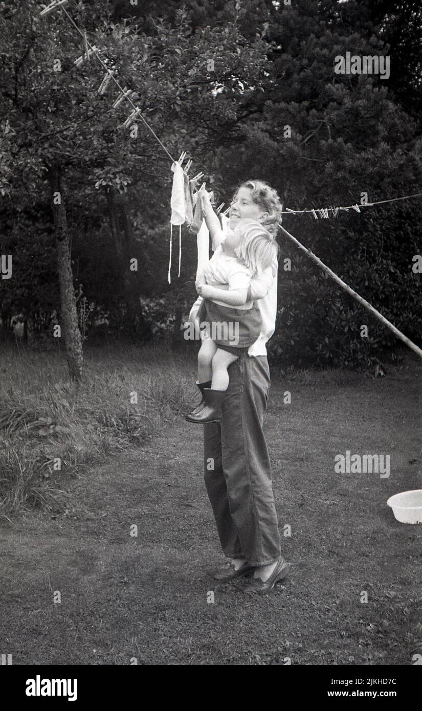 1960s, historical, helping mummy put washing on the clothes line, 'I will do the socks!'....outside in a back garden, a mother holding up her little girl, putting clothes on the line to dry, England, UK. Stock Photo