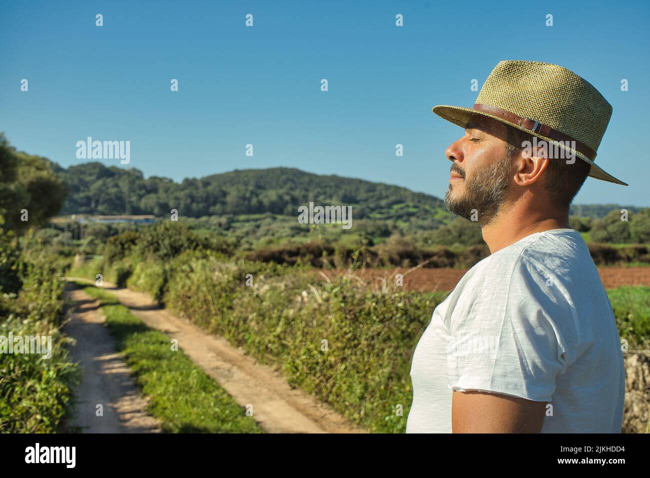 Handsome man in a hat, white t-shirt and jeans relaxes in nature feeling the sun on the skin of his face. Wellness, summertime and vacation concept. Stock Photo