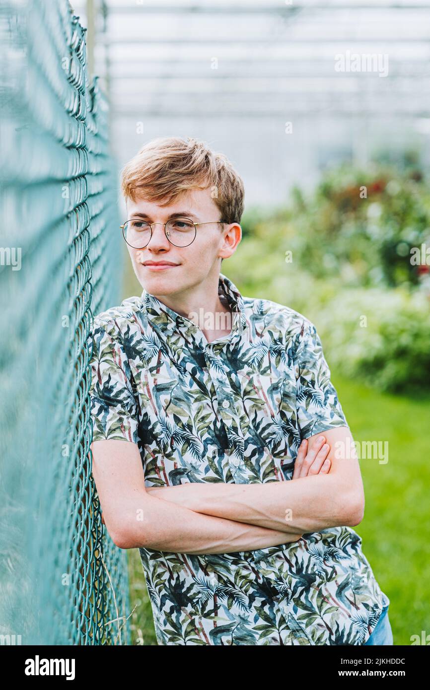 A vertical shot of a young white Caucasian guy with glasses leaning on a fence Stock Photo
