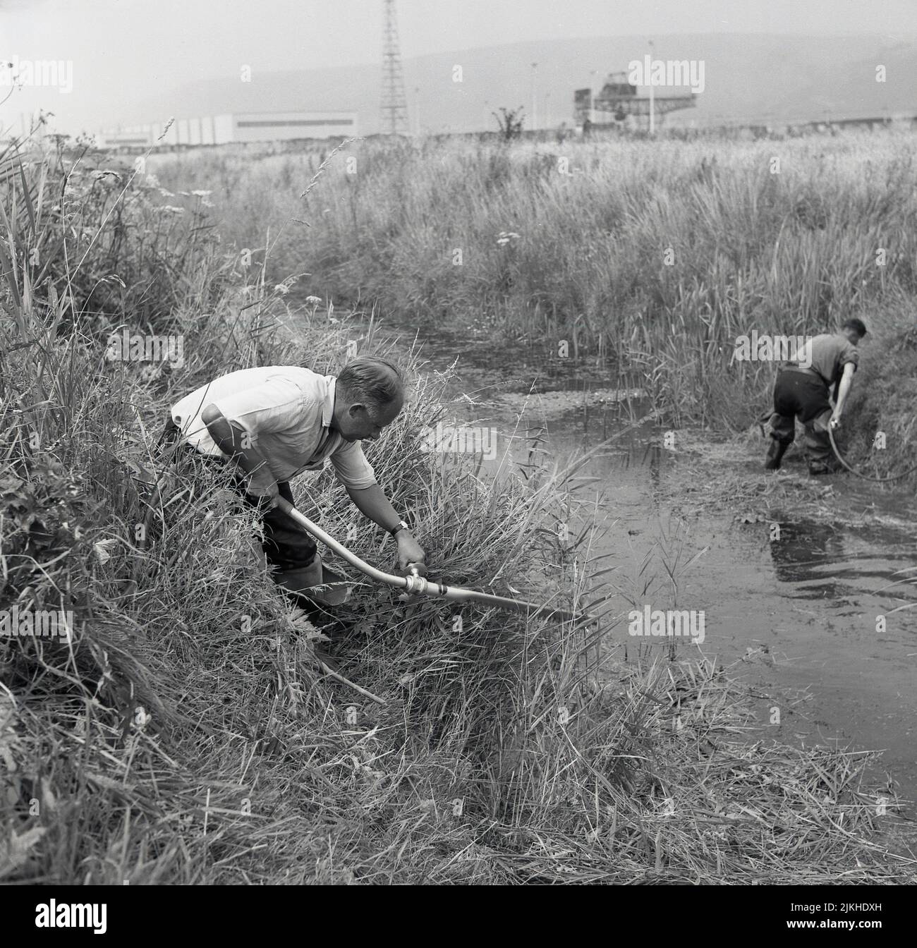 1950s, historical, two workers using hand-held suction apparatus to dredge the bottom of a small river near an industrial site, Port Talbot, Wales, UK. Dredging is a term used for cleaning the bed of a river of mud, silt and other materials, for various reasons, one of which is to improve the flow of water. Stock Photo