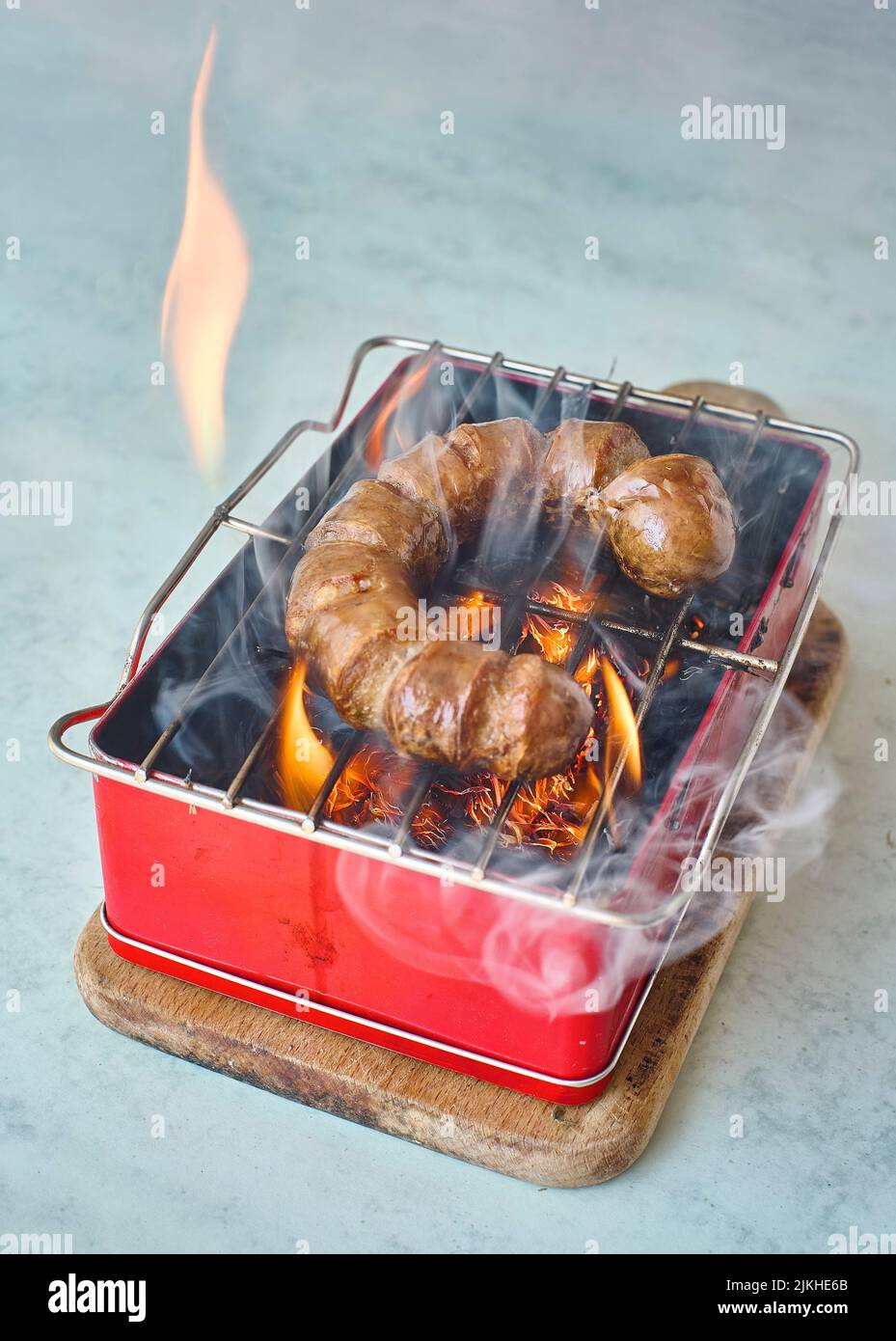 A fresh pork sausage grilling over an improvised barbecue on a metal box. Stock Photo