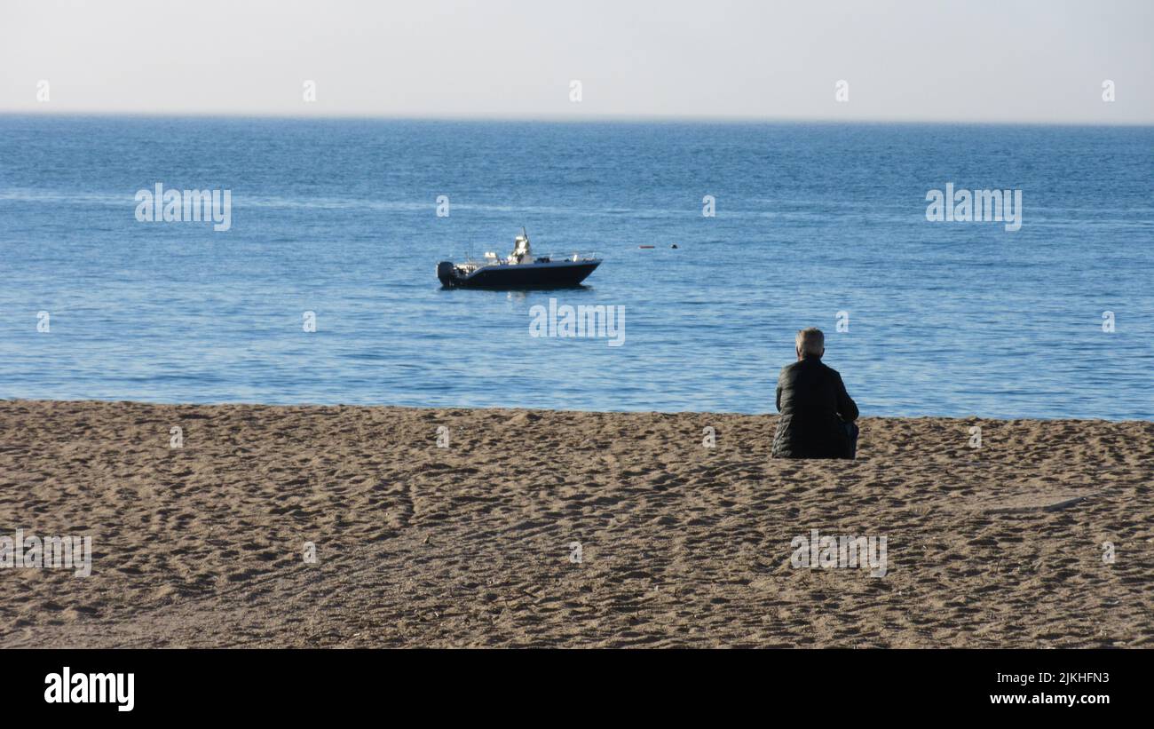 A lonely man sitting at the sandy beach and looking at the sea with a ...