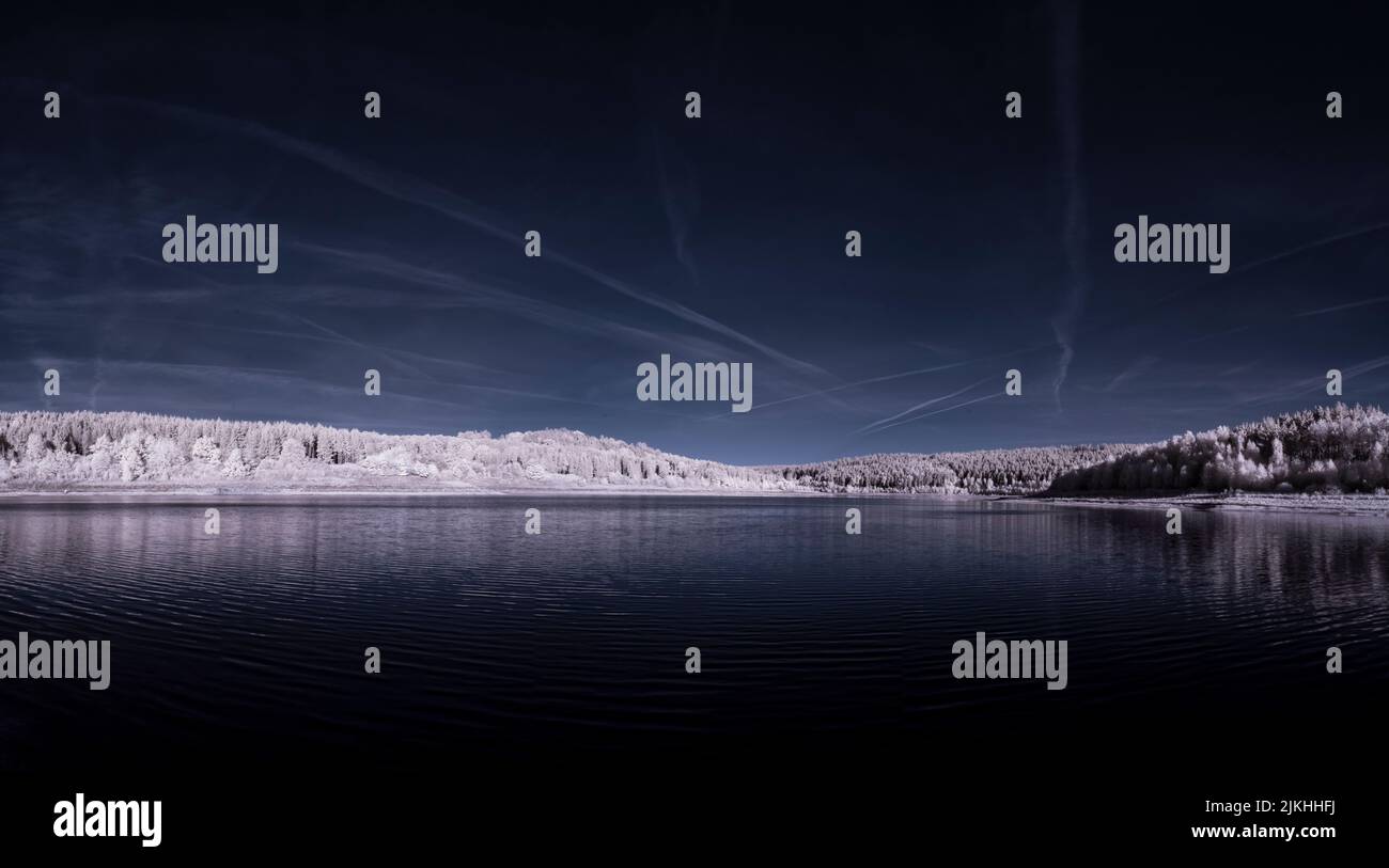 A panoramic view of the blue lake near the snow-covered forest under the dark sky Stock Photo