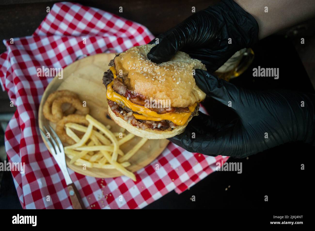 A close-up shot of hands holding a burger in the background of onion rings and french fries on a wooden plate. Stock Photo