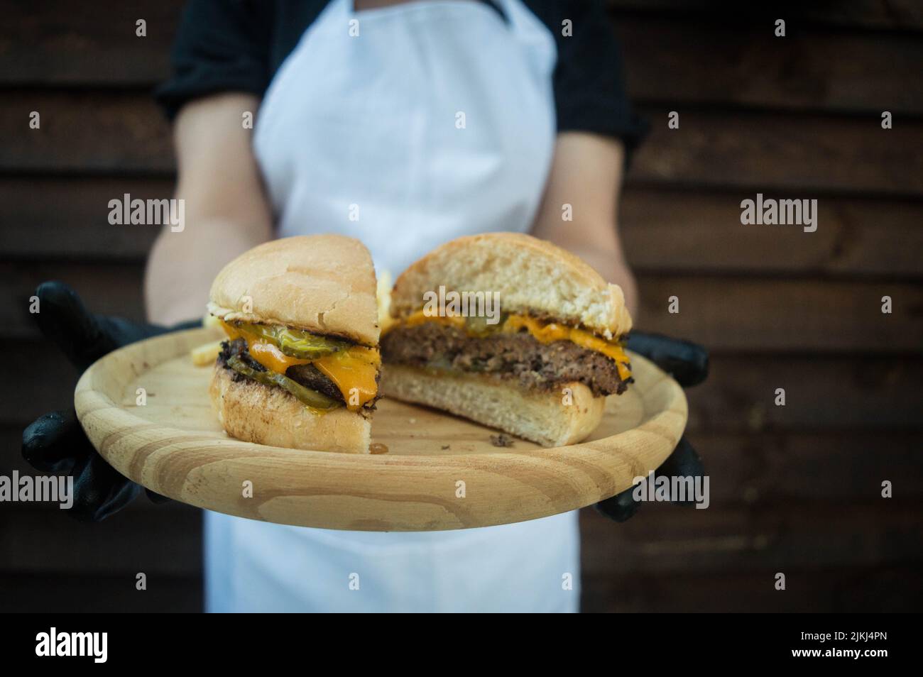 A close-up shot of a chef holding a burger cut in half on a wooden plate. Stock Photo