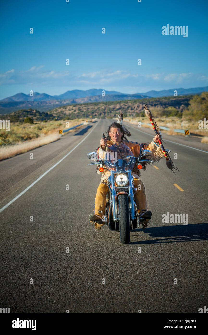 Traditionally dressed Native American man holds coupstick and shoots a gun while driving a Harley Davidson motorcycle down highway in Galisteo Basin, Stock Photo
