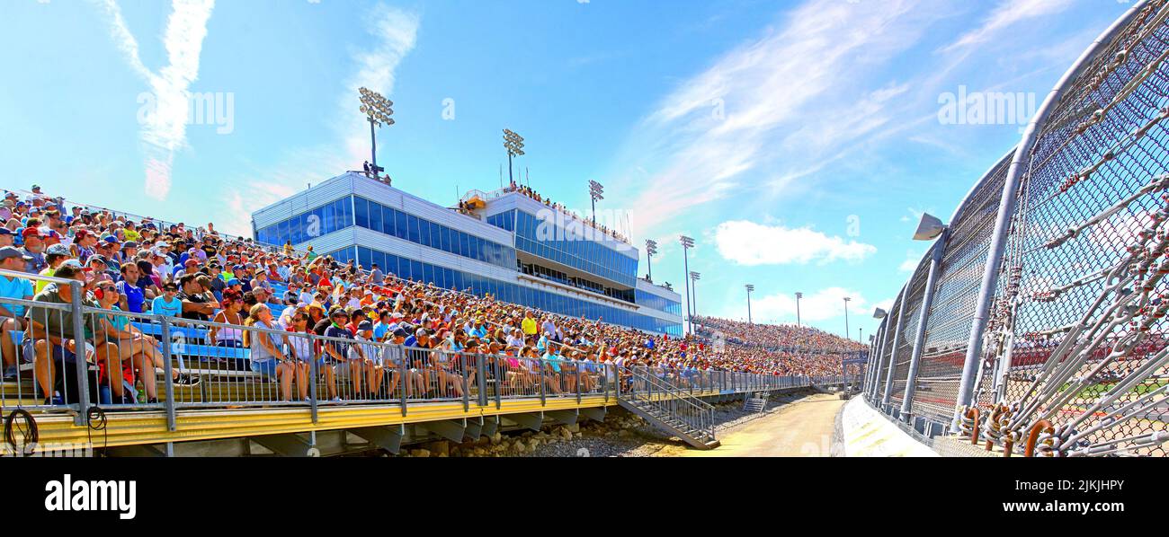 Fans in a grandstand watching a race. Wide view with catch fence and race control tower in the background. Panorama Stock Photo