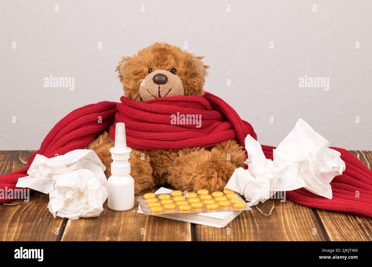 A fluffy teddy bear with a nasal spray, pills, red scarf and tissues on a wooden surface - suffering from illness Stock Photo