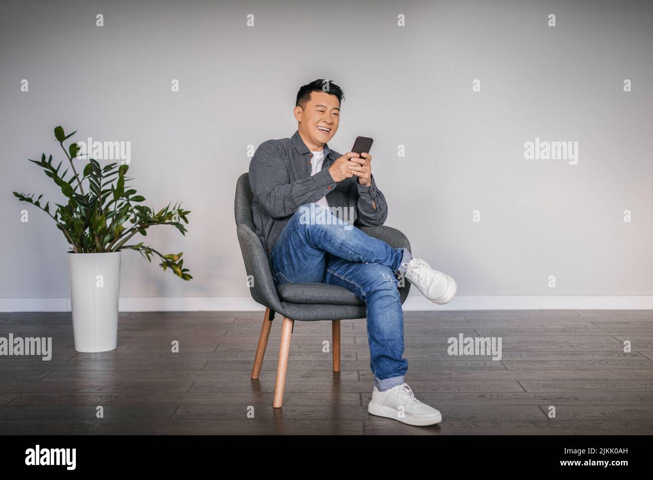 Happy adult asian man typing on smartphone sitting in armchair in minimalist living room interior, free space Stock Photo