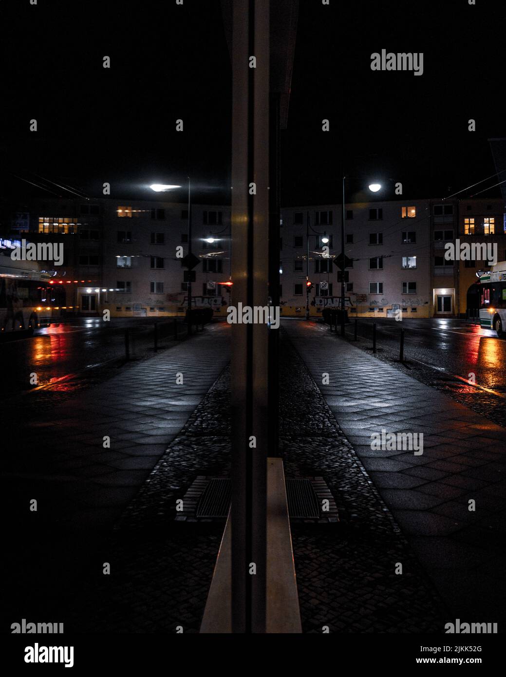 A wet street with tiled floor with shiny light and buildongs with lights under a dark sky at night Stock Photo