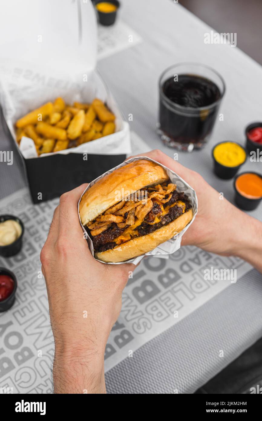 A vertical shot of a person holding a delicious juicy burger and other fast food on a table Stock Photo