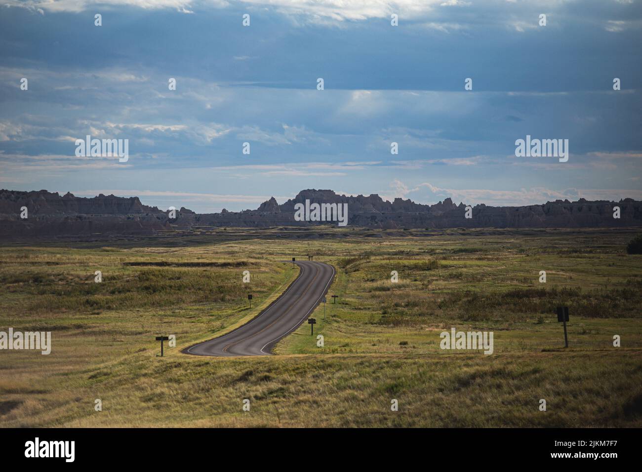 A highway amid green fields on the background of rocky mountains under the blue sky Stock Photo